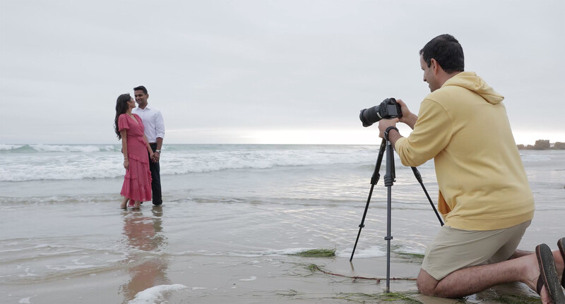 Photographer kneeling on beach sand operating a camera on tripod while couple poses in shallow ocean water.