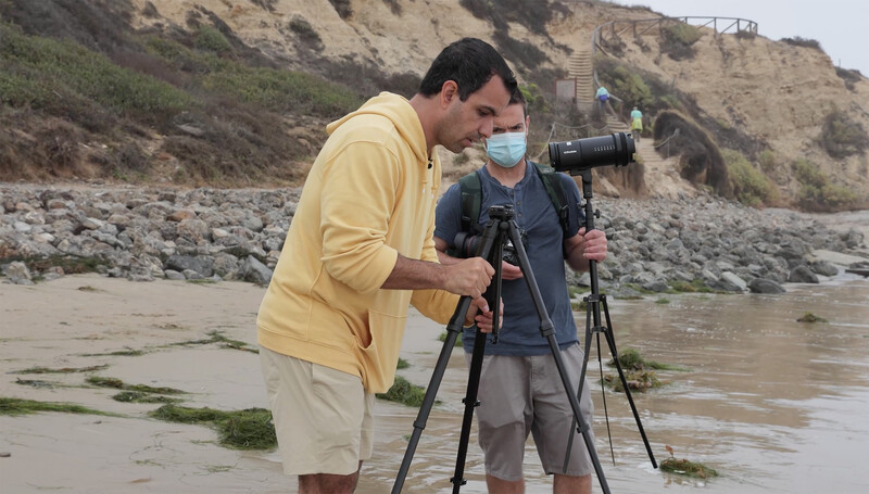 Two people operating a video camera on a tripod at a beach near rocky cliffs and shallow water.