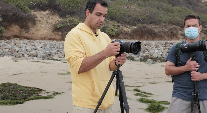 Photographer in yellow hoodie operating camera on tripod at beach location.