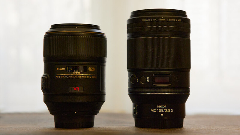 Two Nikon macro lenses side by side on a neutral surface against a blurred background.