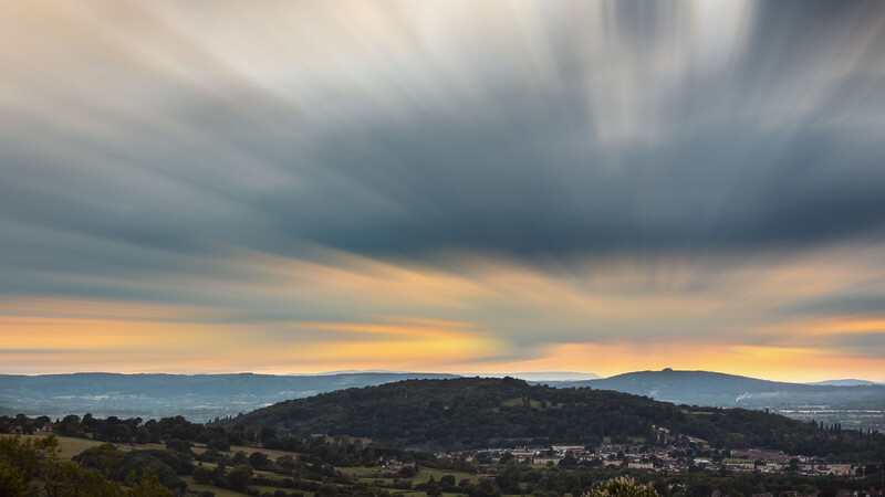Dramatic sunset landscape with layered hills beneath turbulent storm clouds and golden light breaking through.