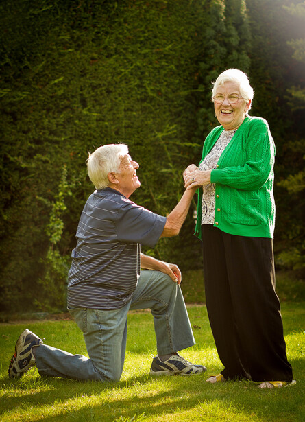 Older man kneeling to propose to an older woman in a grassy garden setting.