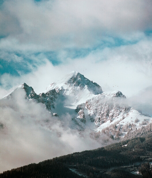 Snow-covered mountain peaks rising through layers of clouds and mist beneath a partly cloudy sky.