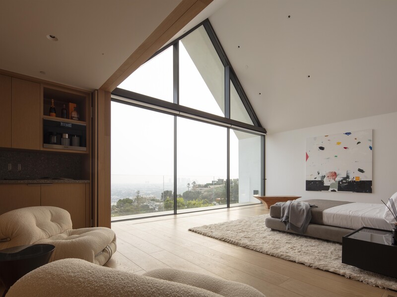 Modern bedroom with floor-to-ceiling triangular windows overlooking a coastal landscape and distant hills.