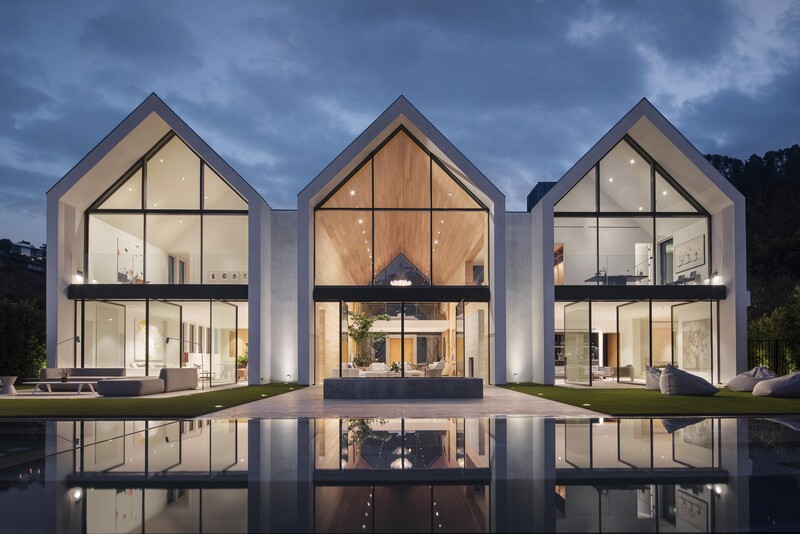 Three modern houses with gabled roofs and large windows reflected in a still pool at dusk.