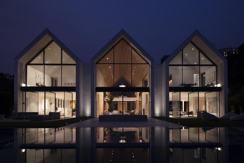 Three illuminated modern houses with peaked roofs reflected in still water at dusk.