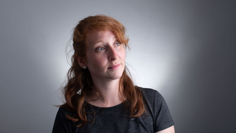 Woman with reddish-brown hair gazing upward against a bright, softly lit gray background.