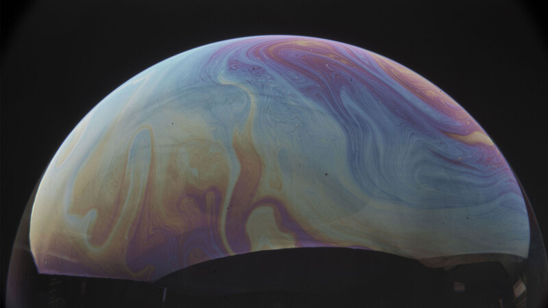 Close-up of a soap bubble surface showing swirling patterns of iridescent colors including purple, blue, and gold against a dark background.