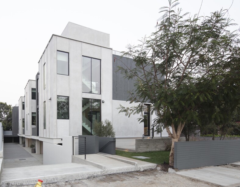 Modern white residential building with clean lines and large windows, flanked by mature trees and metal fencing.
