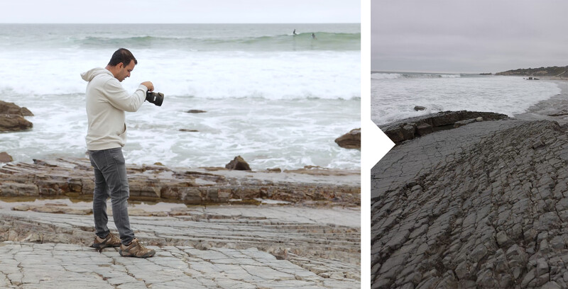 Photographer standing on rocky beach, holding camera to eye while composing a shot of the ocean and coastline.