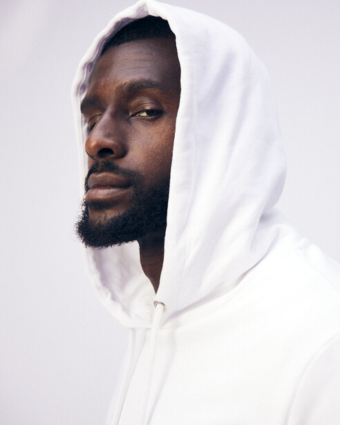 Man wearing white hoodie with hood pulled over head against neutral background.