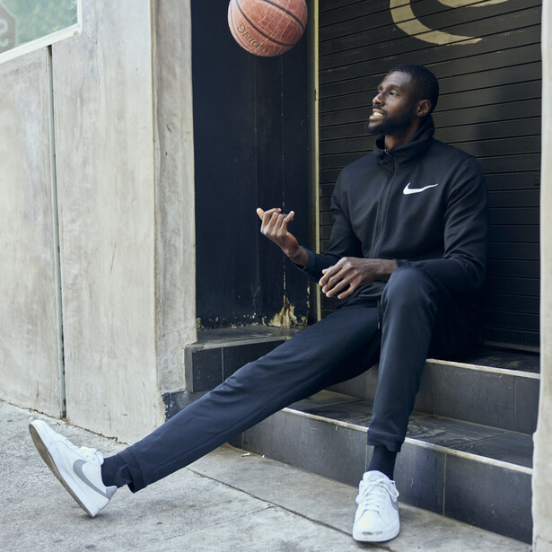 Man in black Nike tracksuit sitting on concrete steps, tossing a basketball upward.