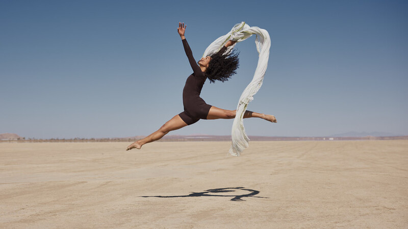 Dancer leaping on a flat desert landscape while holding a flowing white fabric.
