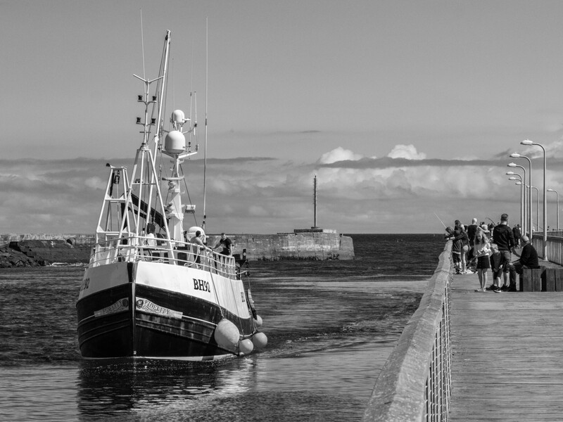 Fishing vessel moored at a pier with people walking on the dock under overcast skies.