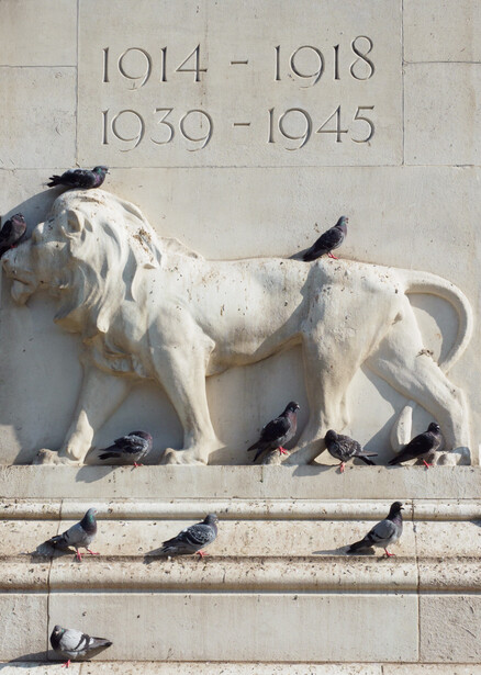 Stone relief sculpture of a reclining lion with birds perched on its body and base, beneath engraved date ranges.