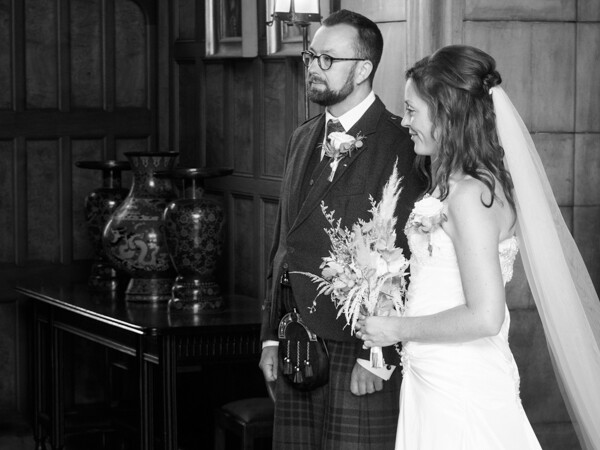 Bride and groom in formal Scottish attire standing indoors near a fireplace in black and white.