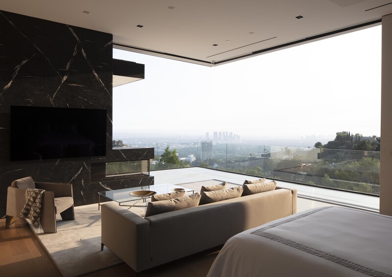 Modern bedroom with floor-to-ceiling windows overlooking a sprawling cityscape at dusk.