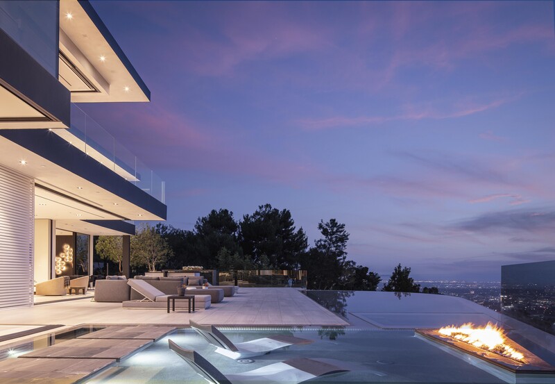 Modern hillside villa at dusk with infinity pool, lounge chairs, and fire pit overlooking city lights.