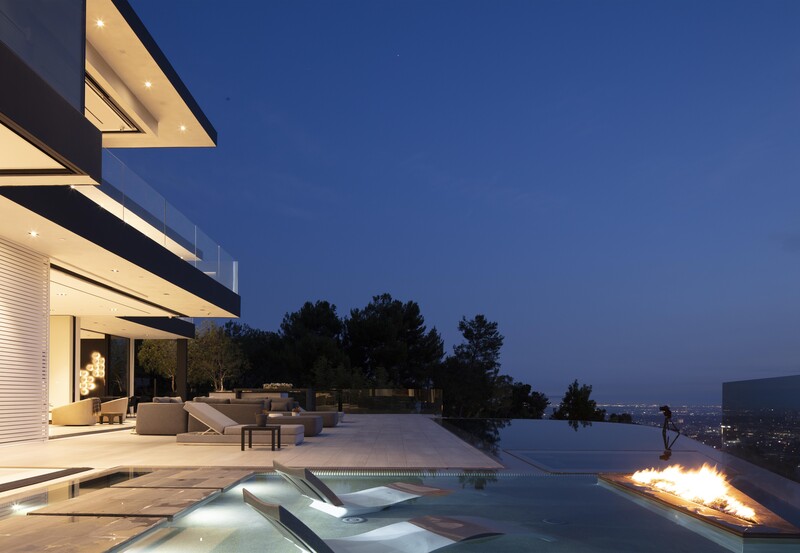 Modern hillside home at dusk with illuminated interior, infinity pool, and fireplace overlooking distant landscape.