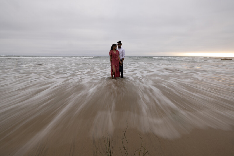 Couple standing in shallow ocean water wearing formal attire as waves blur around them.