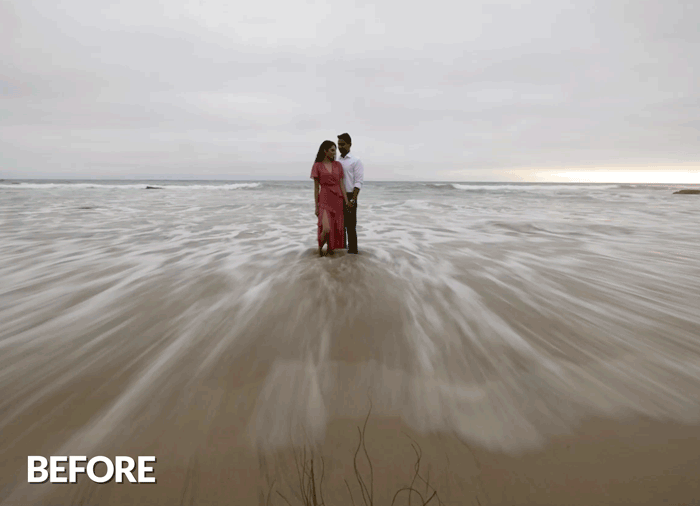 Couple standing in shallow ocean water with motion blur waves in foreground under overcast sky.