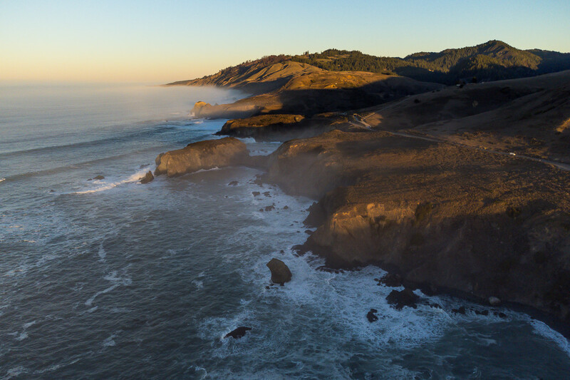 Rocky coastal headland at sunset with golden light illuminating cliffs and dark ocean waves below.