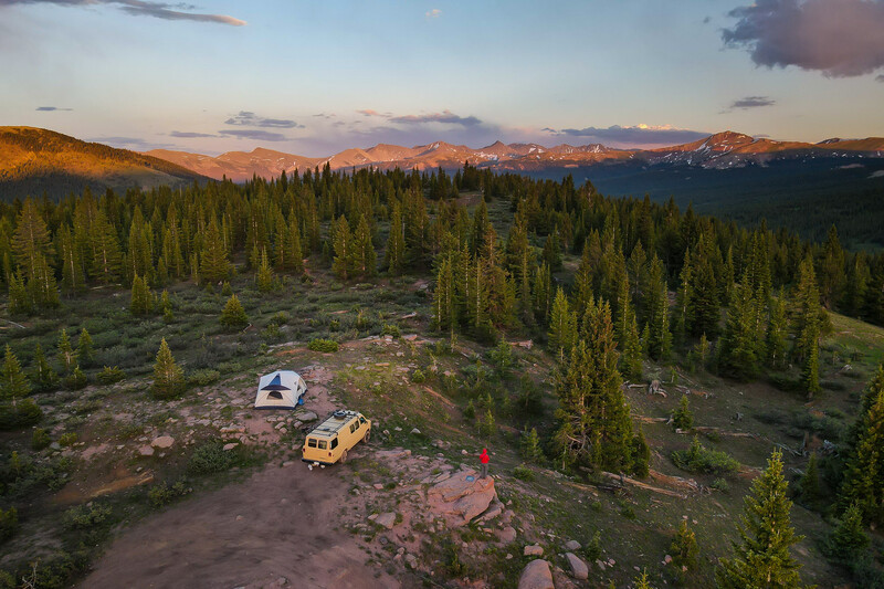Aerial view of a campsite with two vehicles among coniferous forest at sunset, mountains visible in distance.