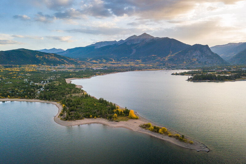 Aerial view of a pristine alpine lake surrounded by forested shores, mountains, and scattered settlements under dramatic sky.