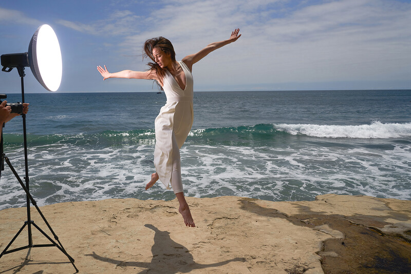 Woman in white outfit jumping on beach with studio lighting rig visible.