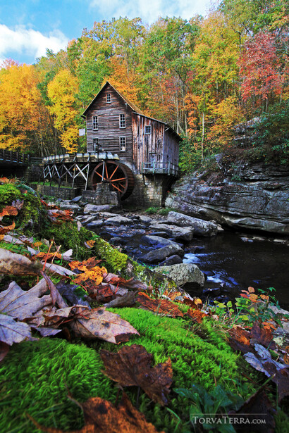 Historic wooden mill building with large water wheel beside flowing stream surrounded by autumn foliage.