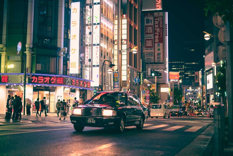 Black taxi cab crossing a zebra crossing in a neon-lit urban street at night surrounded by pedestrians and illuminated storefronts.