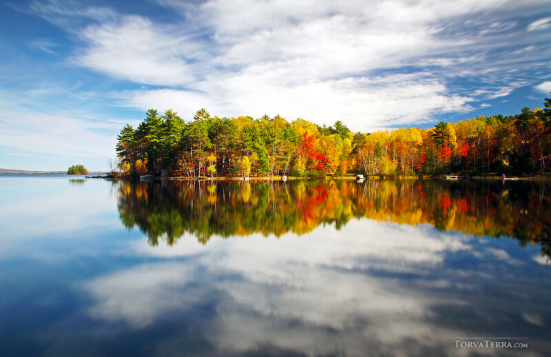 Autumn forest reflected in still lake water under partly cloudy sky.