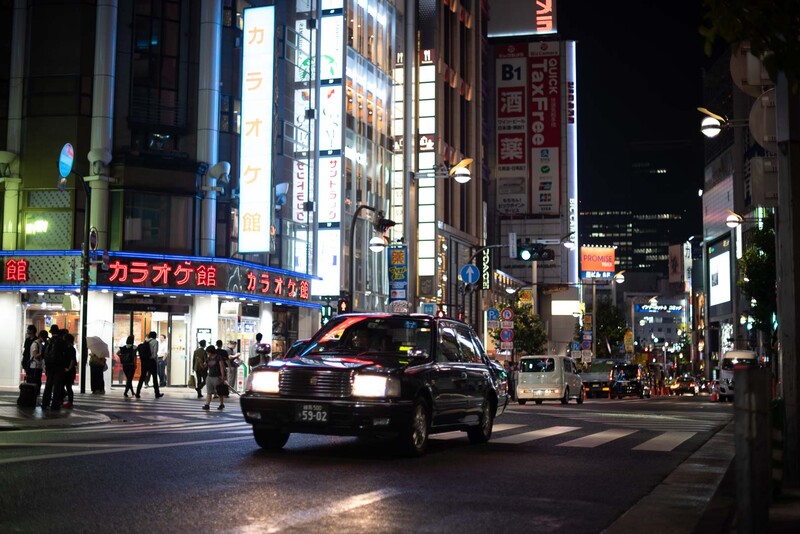Nighttime urban street scene with taxi and pedestrians under neon signs.