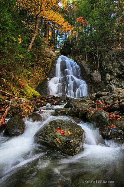 Multi-tiered waterfall cascading over moss-covered rocks through autumn forest.