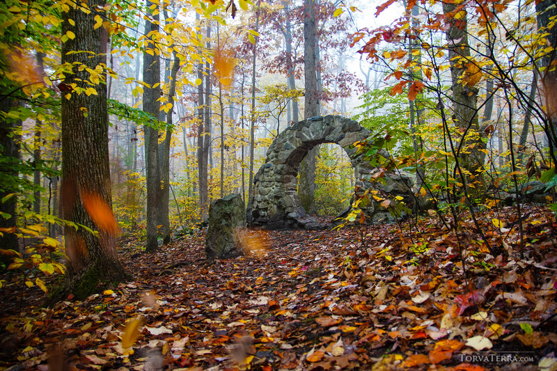 Stone arch structure in autumn forest surrounded by fall foliage and bare trees.