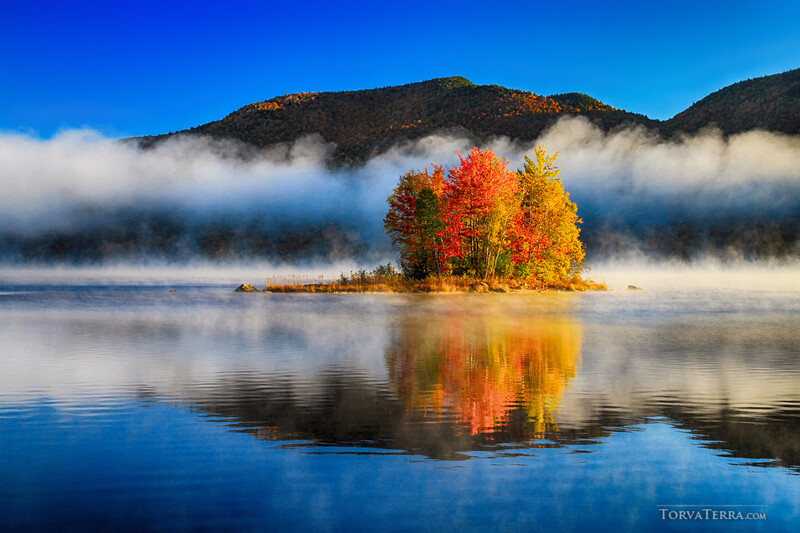 Small forested island with vibrant autumn foliage reflected in still water, surrounded by mist-covered mountains under clear blue sky.