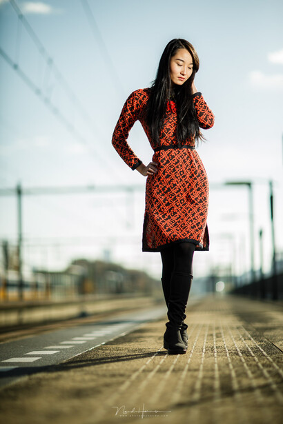 Woman in red patterned dress standing on railroad tracks with overcast sky behind her.