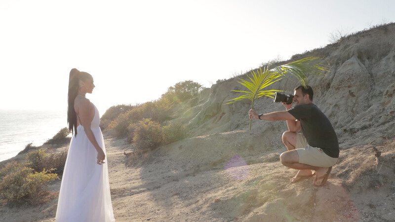 Photographer crouching with camera while woman in white dress poses on sandy coastal bluff.