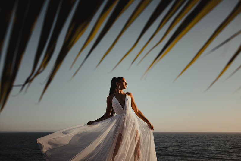 Woman in flowing white gown standing at ocean's edge at sunset, with draped fabric overhead creating diagonal line patterns.