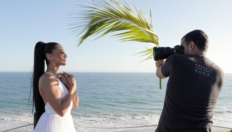 Photographer shooting a portrait of a woman in a white dress on a beach, with a palm frond in the foreground.
