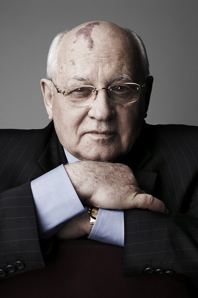 Older man with glasses wearing a dark suit and light blue dress shirt, posed with hands clasped at chest against neutral gray background.