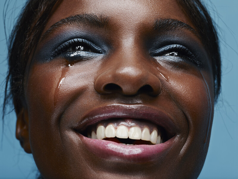 Close-up portrait of a woman with dark eyeshadow and white teeth smiling broadly against a blue background.
