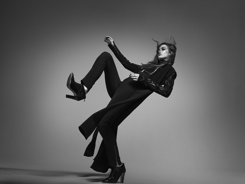 Person in black suit and heeled boots posed dynamically against a neutral background in a black and white studio photograph.