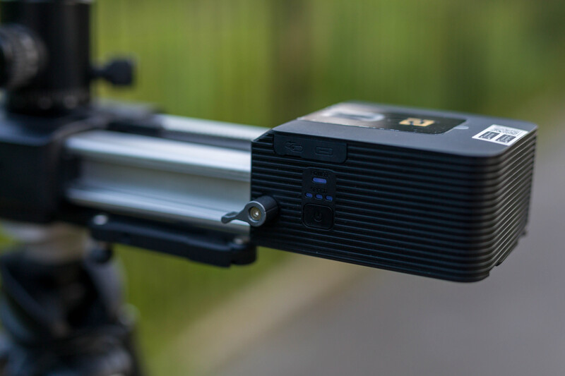 Compact black projector mounted on a tripod with a silver extension arm against a blurred green background.