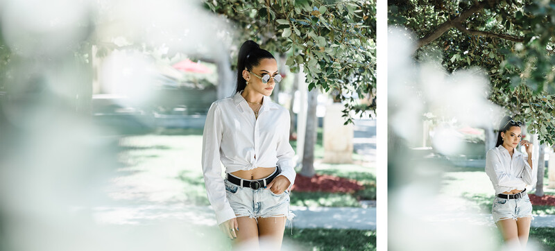 Woman in white shirt and denim shorts standing under tree canopy in bright sunlight.