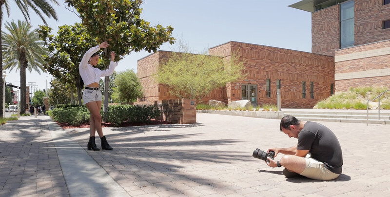 Photographer crouching low while shooting a model posing against brick architecture in bright daylight.