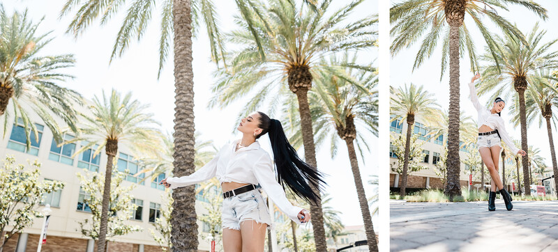 Woman in white shirt and denim shorts posing among palm trees in a sunny urban plaza.