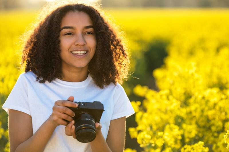 Smiling photographer holding a DSLR camera in a bright yellow flower field.