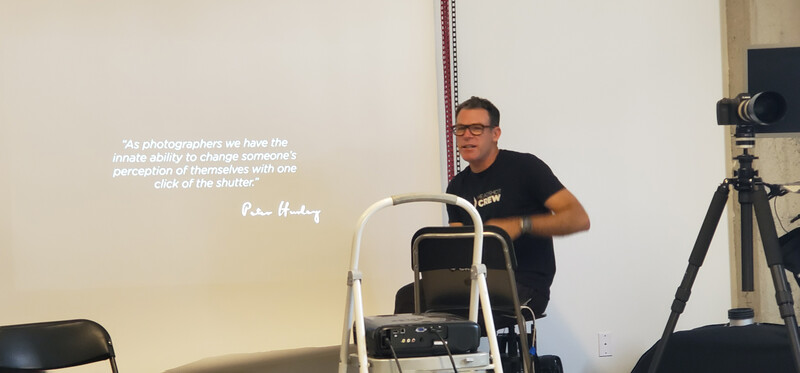 Photographer in black shirt standing next to tripod-mounted camera during presentation with quote about photography on wall behind.
