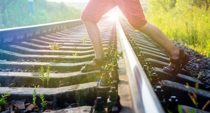 Person's legs straddling railroad tracks with green grass and bright sunlight in the background.